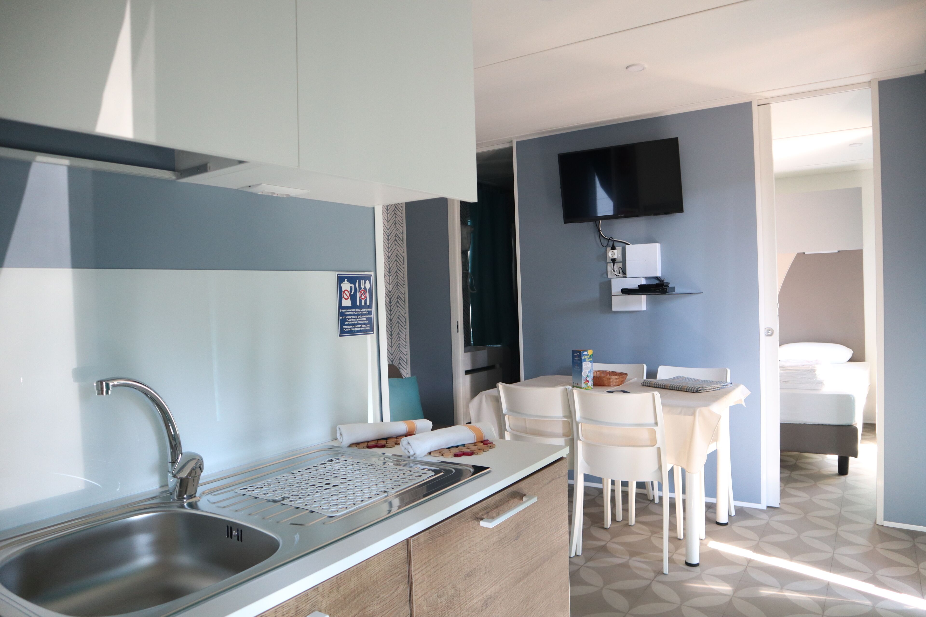 A modern kitchen with a stainless steel sink, a dining area with white chairs and a table, a flat-screen TV mounted on the wall, and a bed in the adjacent room.