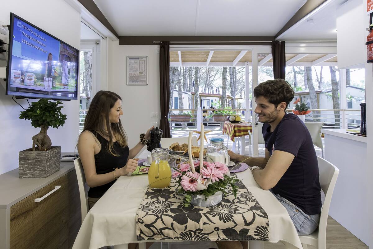 Een eethoek met een tafel gedekt voor twee personen, een aan de muur bevestigde televisie en een uitzicht op de omgeving door grote ramen.