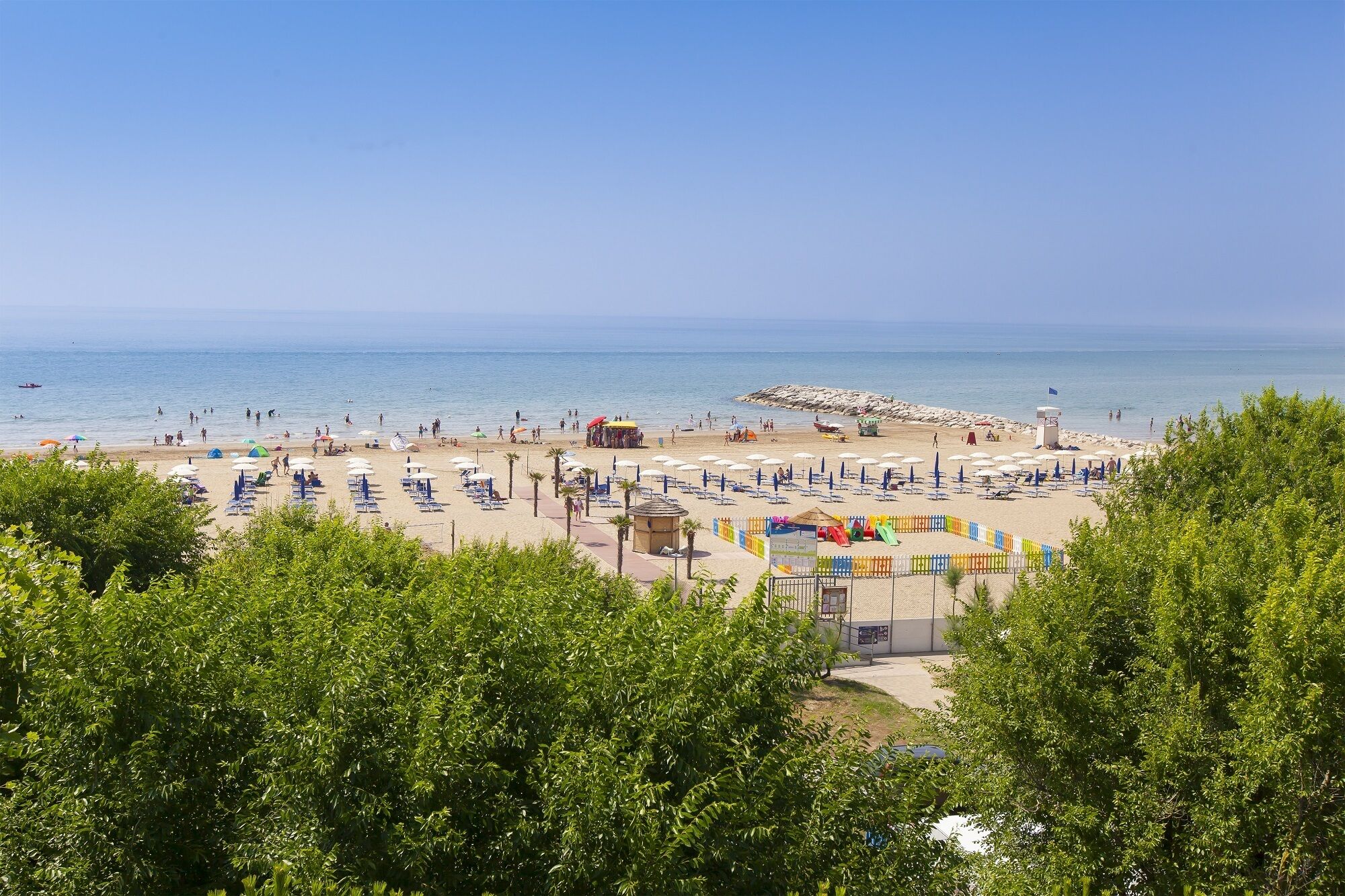A beach with rows of sun loungers and umbrellas, a lifeguard tower, and a rocky breakwater.