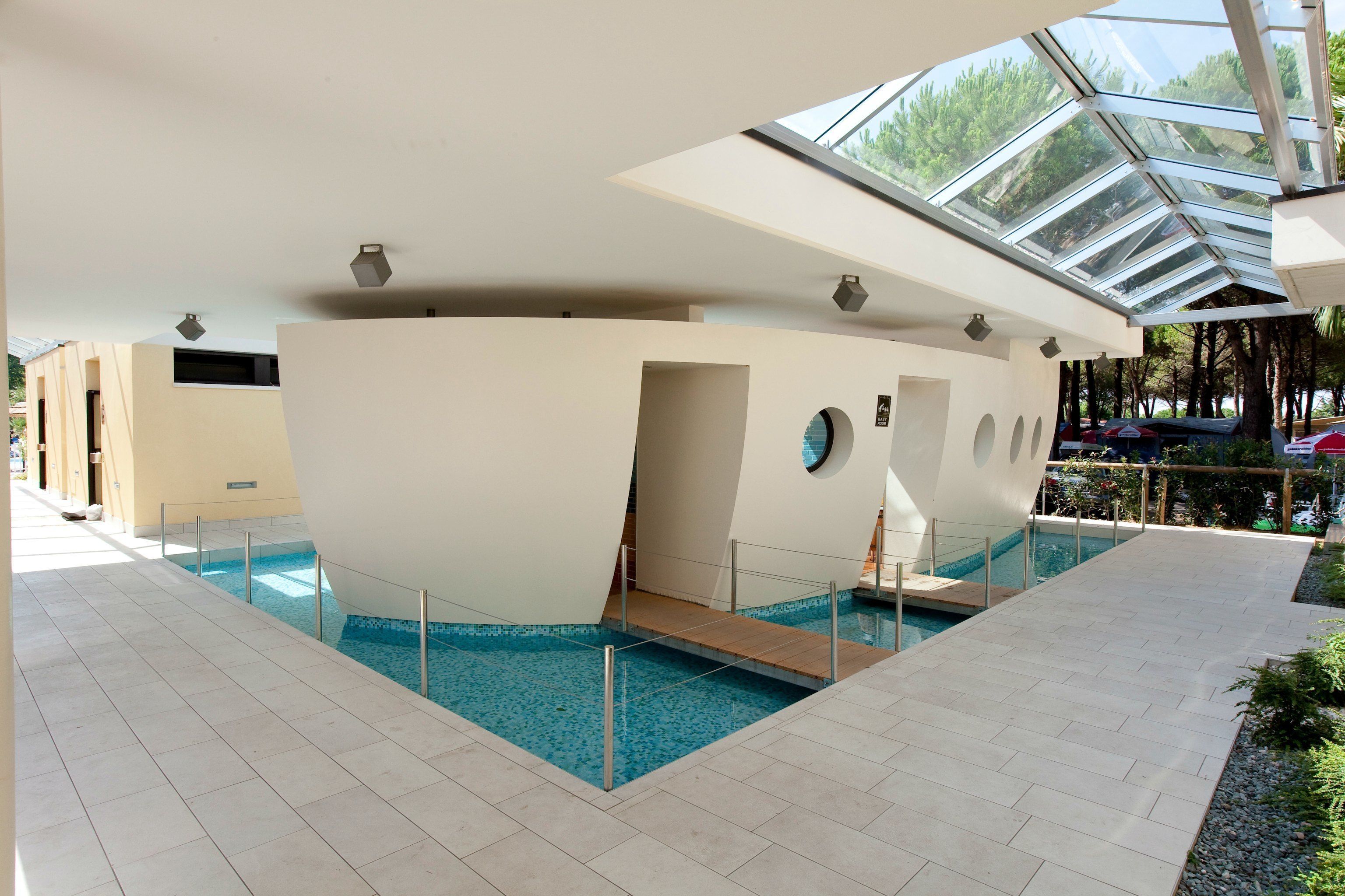 Une piscine intérieure moderne dotée d’un toit en verre, entourée d’une allée carrelée et d’une petite terrasse en bois.