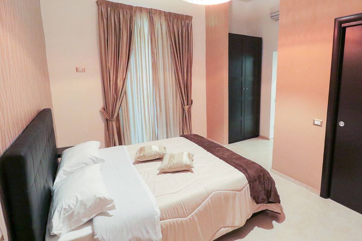 A neatly made bed with pillows and a blanket, a dark headboard, a large window with curtains, and a door to the right.