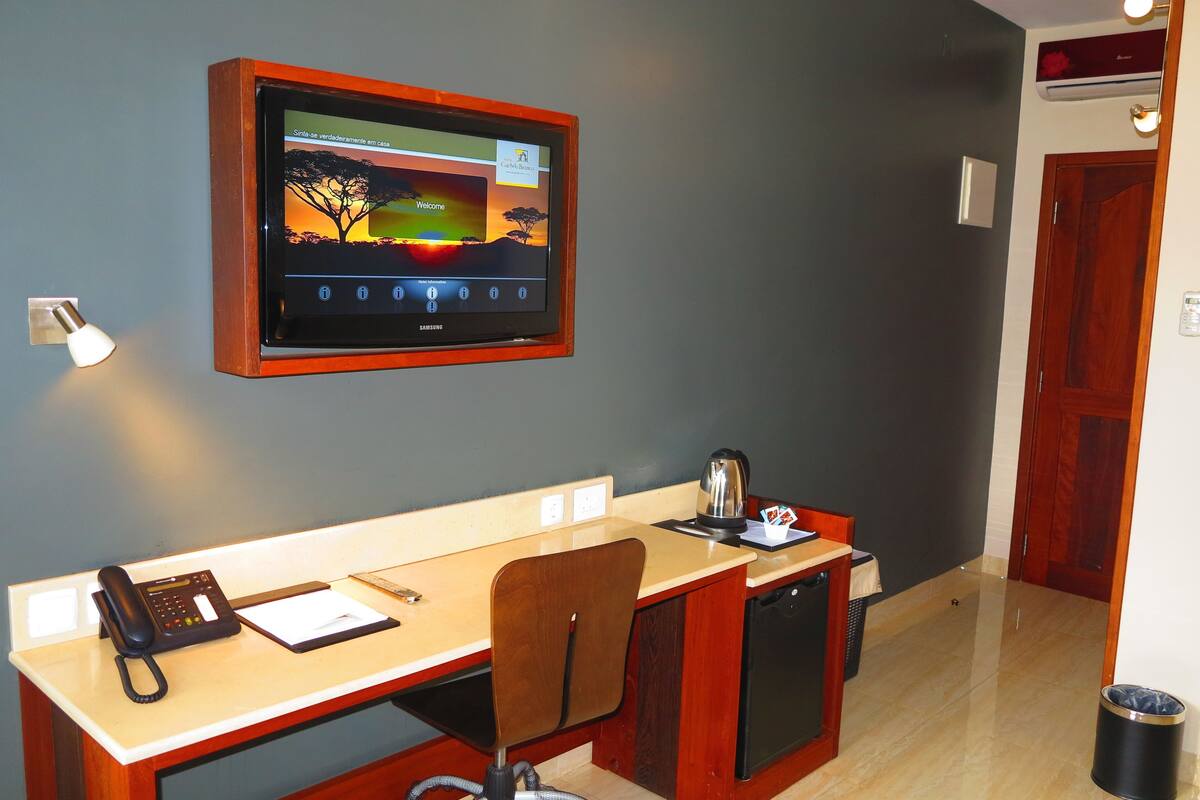 A hotel room with a desk, chair, a small kitchenette, a television mounted on the wall, and a telephone.