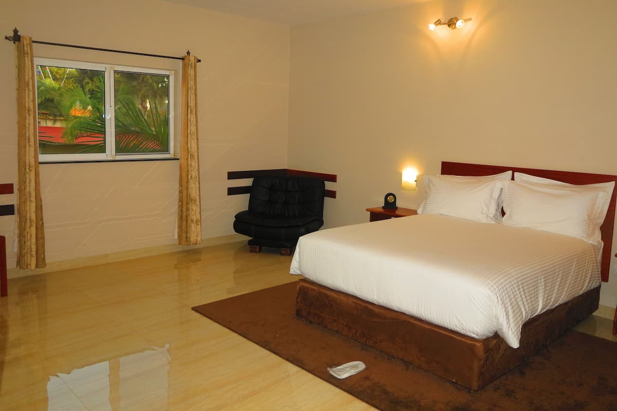 A bedroom with a bed, a chair, a nightstand, a wall-mounted light, and a window with curtains.