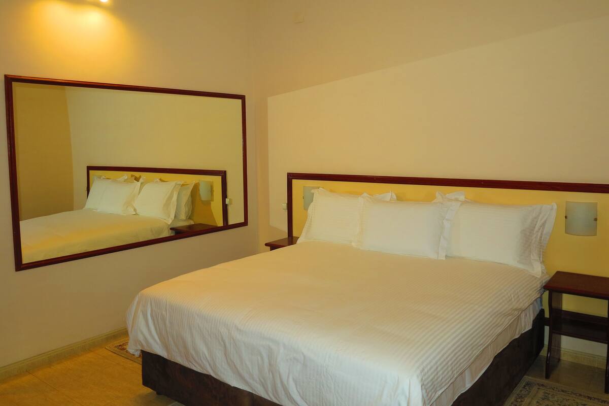 A hotel room with a large bed, a bedside table, a nightstand, and a wall-mounted mirror.