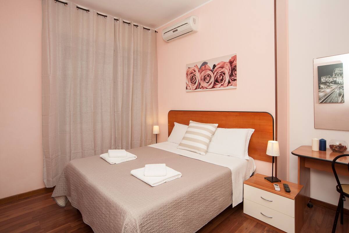 A hotel room with a bed, a nightstand, a lamp, and a wall-mounted air conditioner.