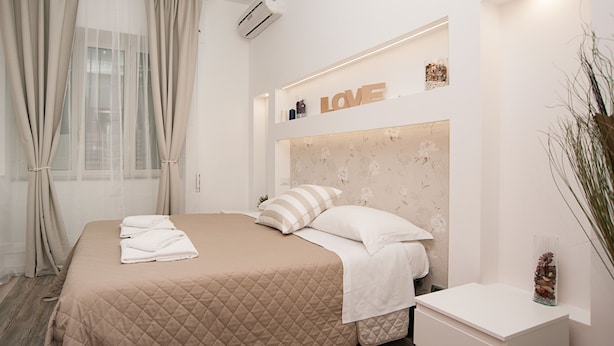 Une chambre d’hôtel moderne avec un grand lit, des rideaux beiges, un climatiseur fixé au mur et une étagère décorative sur laquelle est inscrit le mot « AMOUR ».
