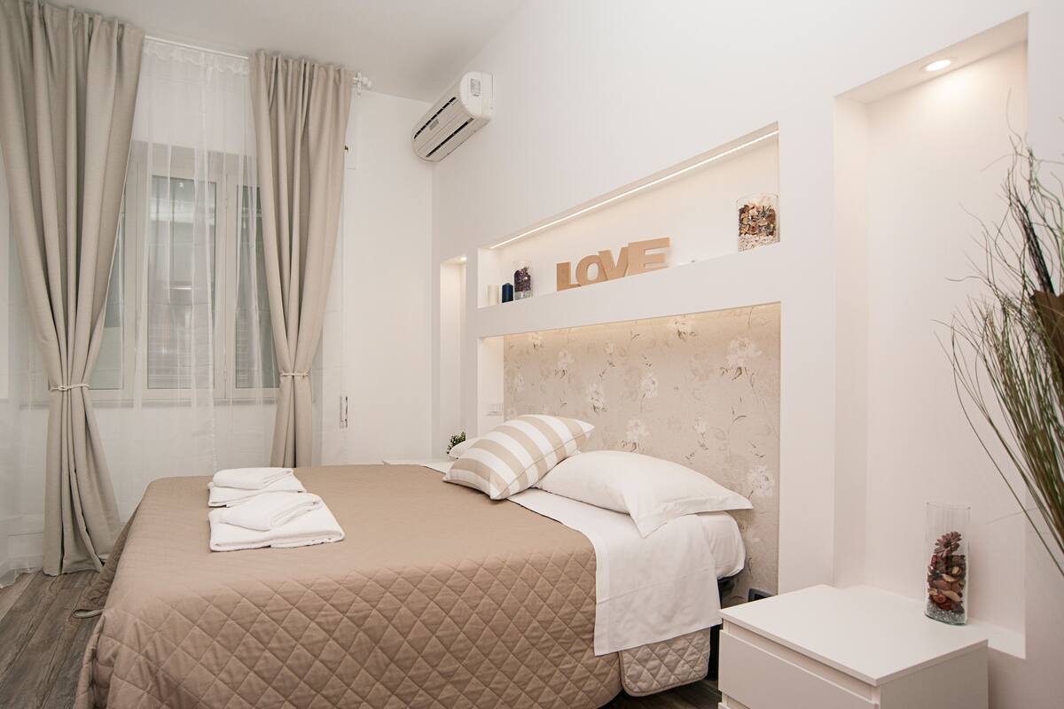A modern hotel room with a large bed, beige curtains, a wall-mounted air conditioner, and a decorative shelf with the word "LOVE".