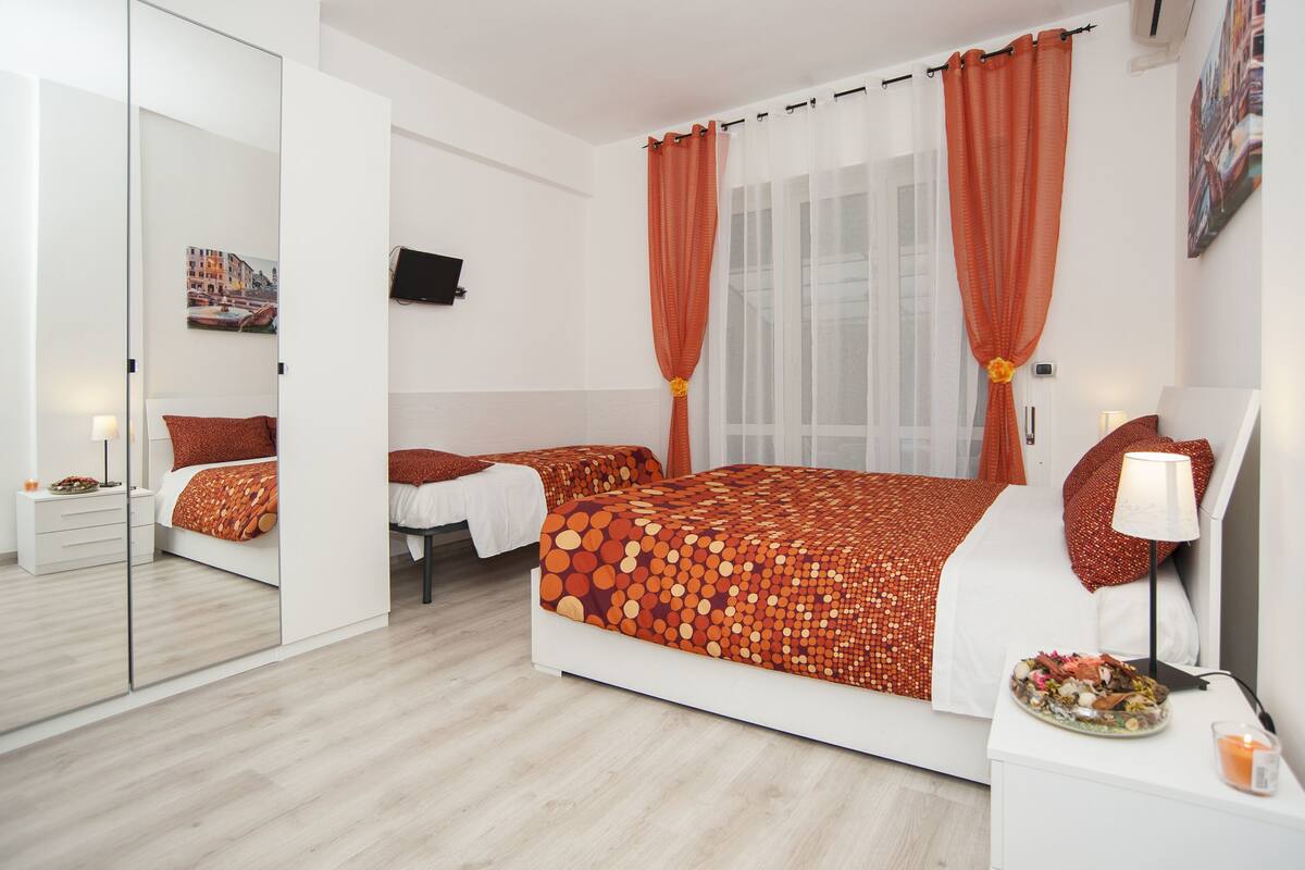 A modern bedroom with a large bed, a smaller bed, a bedside table with a lamp, a mirror, and a wall-mounted TV.