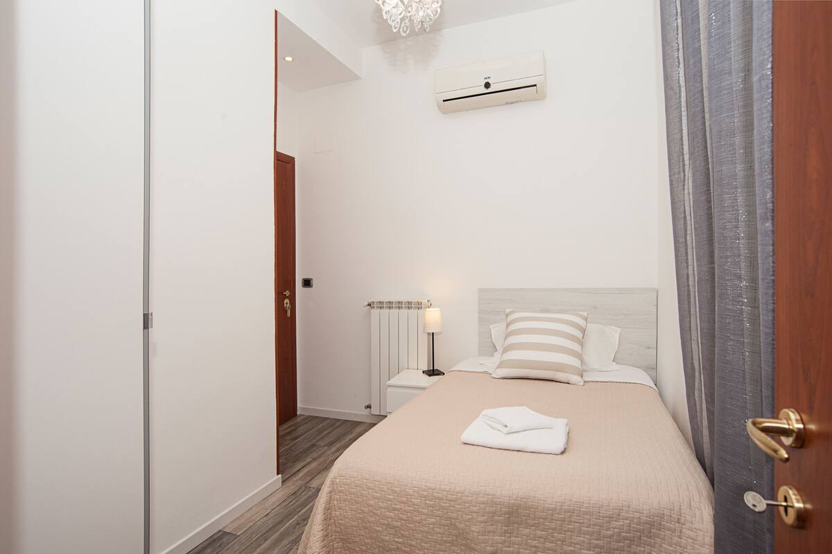 A hotel room with a bed, a nightstand, a lamp, a radiator, and an air conditioning unit.