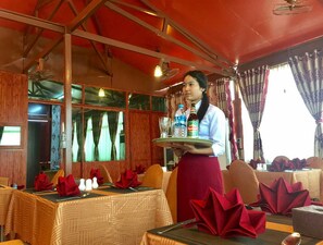 A restaurant interior with wooden paneling, tables set with red napkins, and a server holding a tray with bottles. - Bagan Mingalar Hotel (Mandalay)