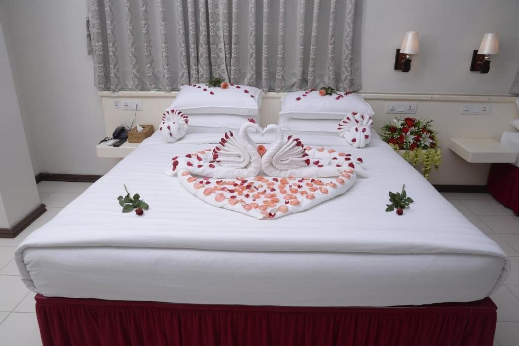 A neatly made bed with white linens, a heart-shaped towel arrangement, and rose petals.