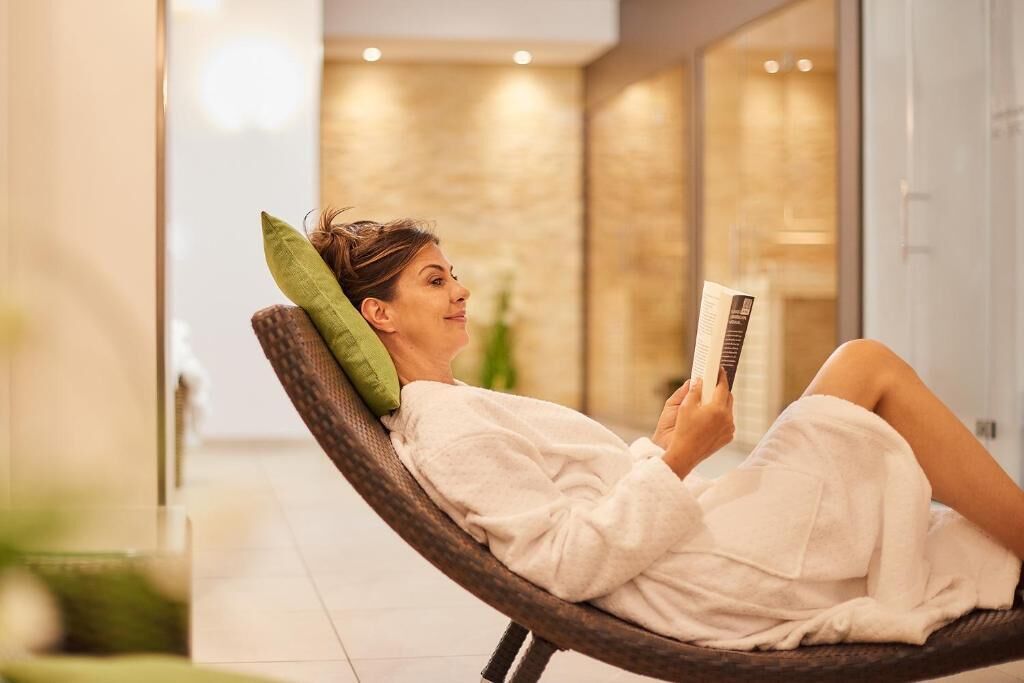 A person reclining in a wicker chair with a green pillow, reading a book in a well-lit indoor space with a tiled floor and a glass wall.