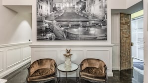 En entréhall med ett stort svartvitt fotografi av en storslagen trappa och en fontän, två bekväma fåtöljer, ett litet bord med en dekorativ prydnad och en dörr med en vägg som har en accentfärg i tegel.