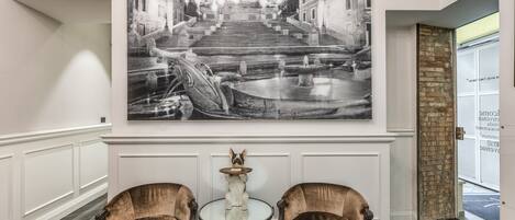 A lobby with a large black and white photograph of a grand staircase and fountain, two plush armchairs, a small table with a decorative item, and a door with a brick accent wall.