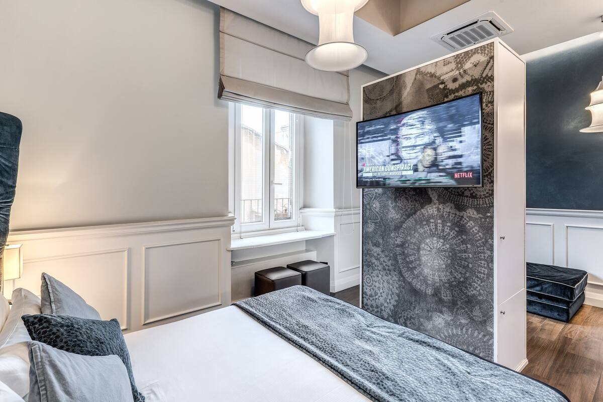 A modern hotel room with a large bed, a TV mounted on a textured wall, a window with a view, and a small seating area.