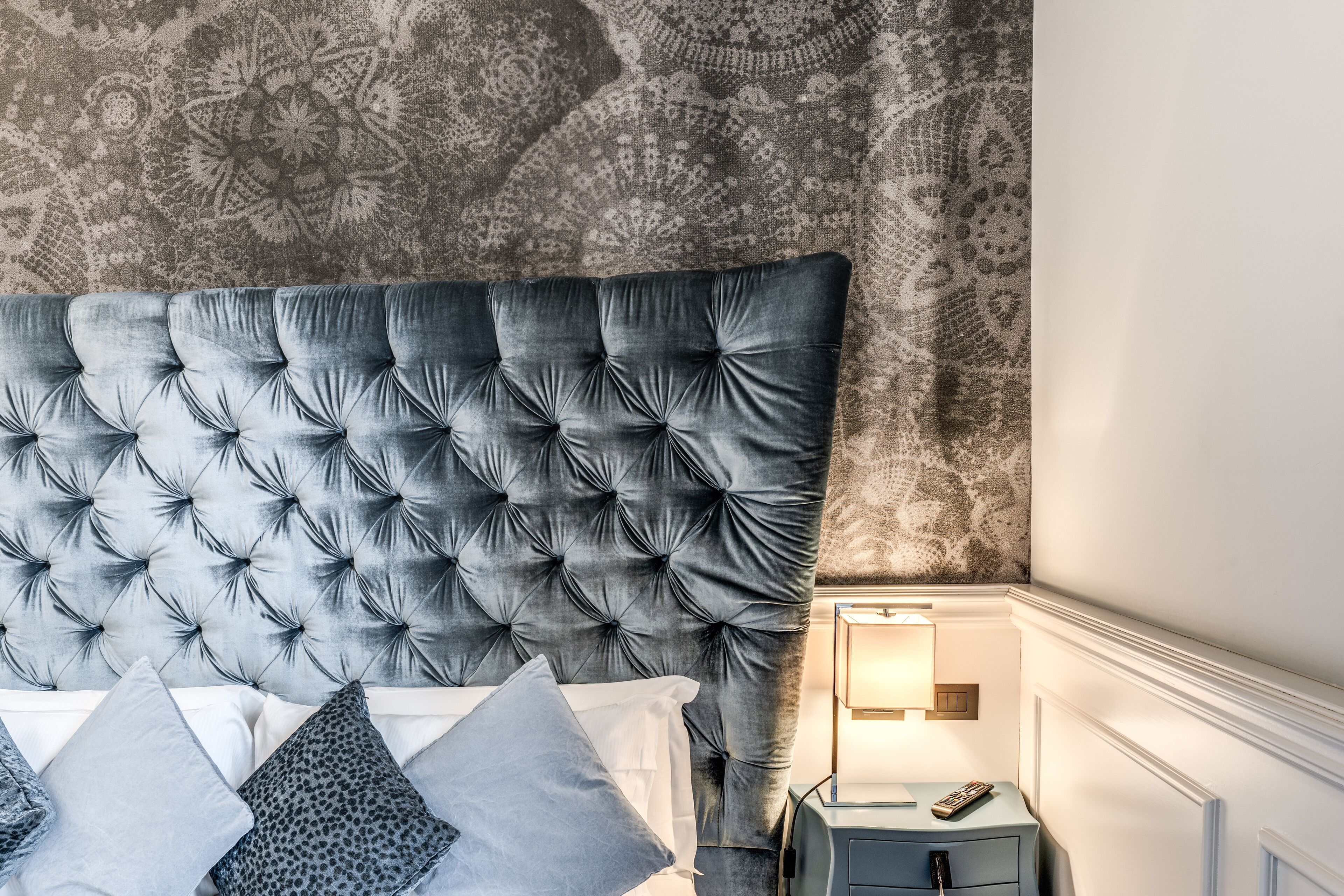 A bedroom with a tufted headboard, a patterned wall, and a bedside table with a lamp.