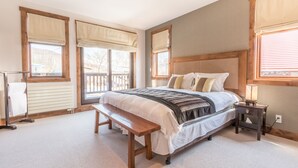 A bedroom with a large bed, wooden headboard, and a bench. There are windows with curtains, a nightstand with a lamp, and a radiator. - Yukimi 1 (Kutchan)
