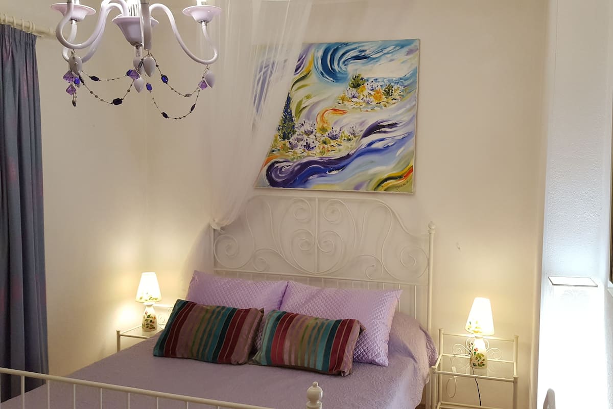 A bedroom with a bed, two bedside lamps, a chandelier, and two paintings on the walls.