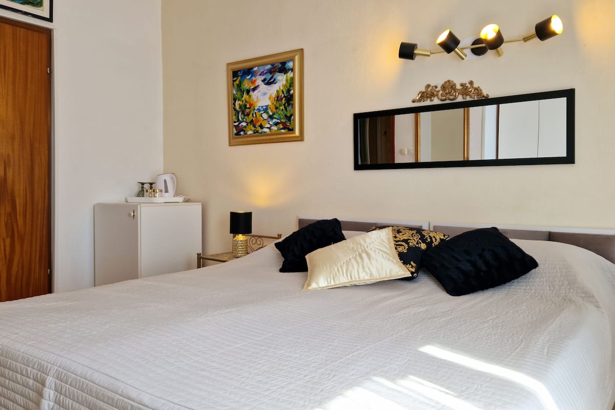 A hotel room with a large bed, a bedside table with a lamp, a small cabinet, a painting on the wall, and a mirror above the bed.