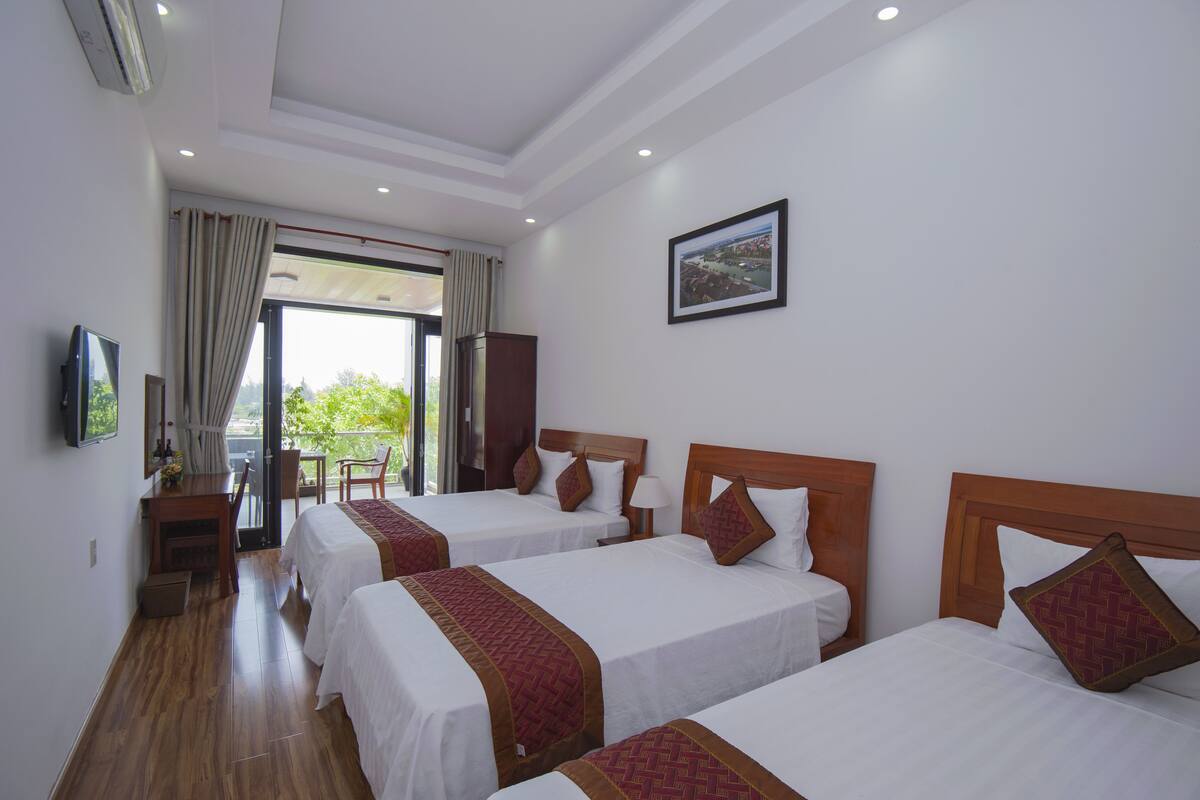 A hotel room with three beds, a desk with a chair, a TV, and a large window with curtains.