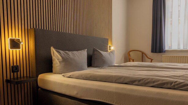 A modern bedroom with a large bed, a headboard with integrated lighting, a bedside table, and a window with curtains. - Hotel Rendsburg (Rendsburg)