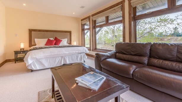 Un dormitorio con una cama grande, un sofá de cuero marrón, una mesa de centro de madera y vistas a árboles a través de amplios ventanales.