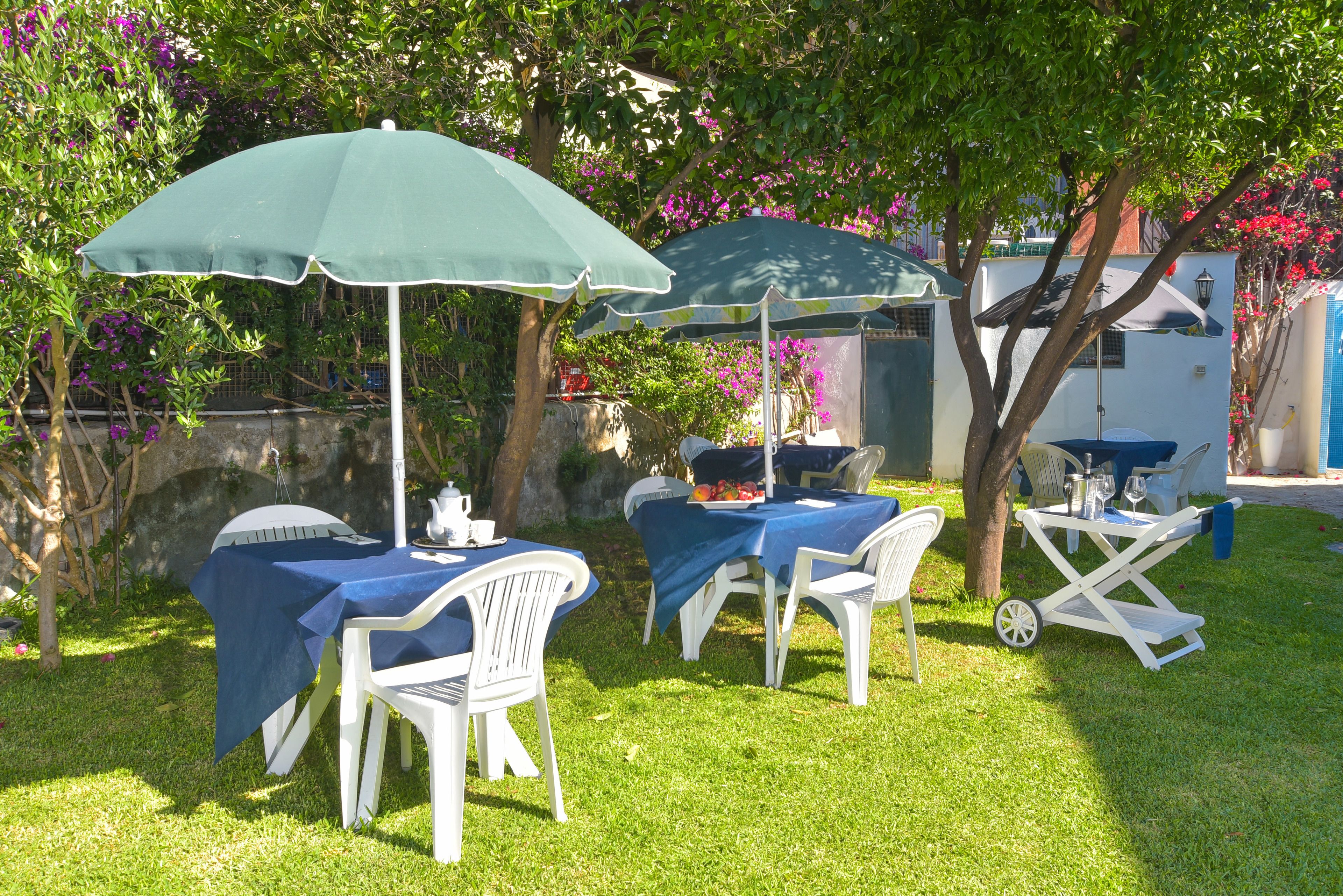 An outdoor patio with tables and chairs, covered by umbrellas, set against a backdrop of trees and a building.