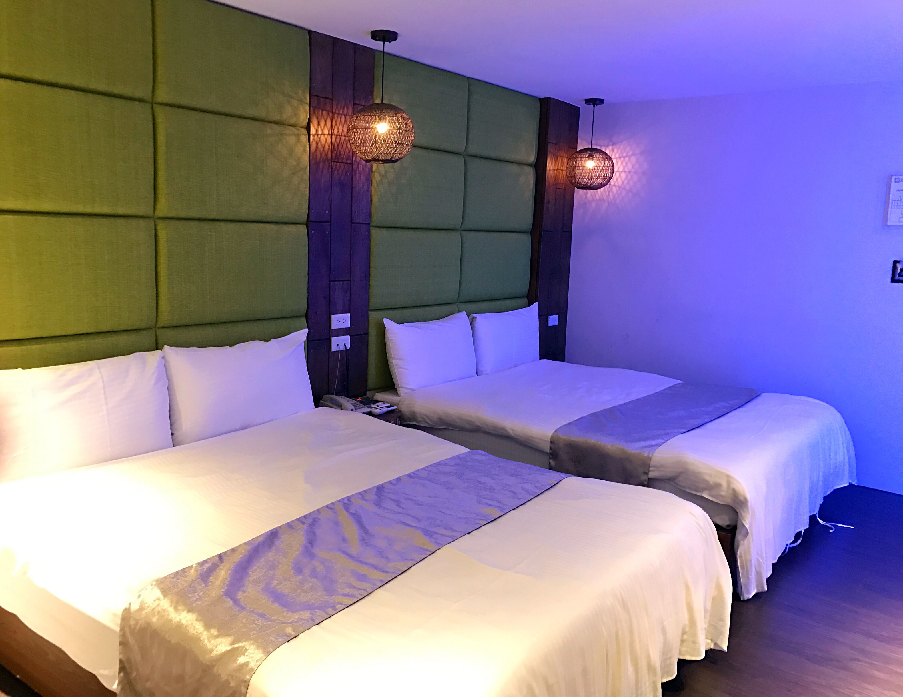 A hotel room with two beds, a green upholstered headboard, and two wall-mounted pendant lights.