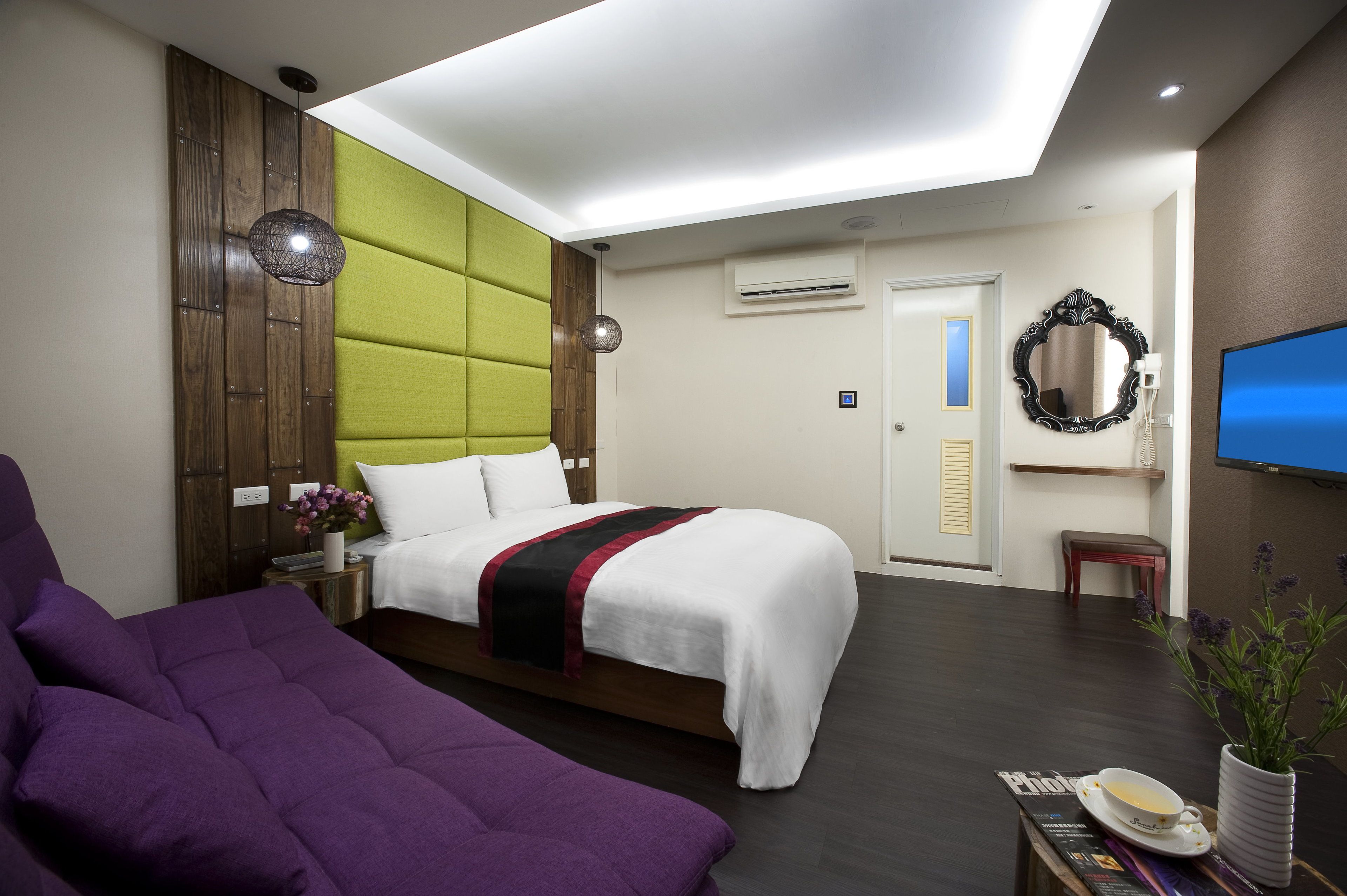 A modern hotel room with a bed, a purple sofa, a small table with a cup and a vase, and a television.