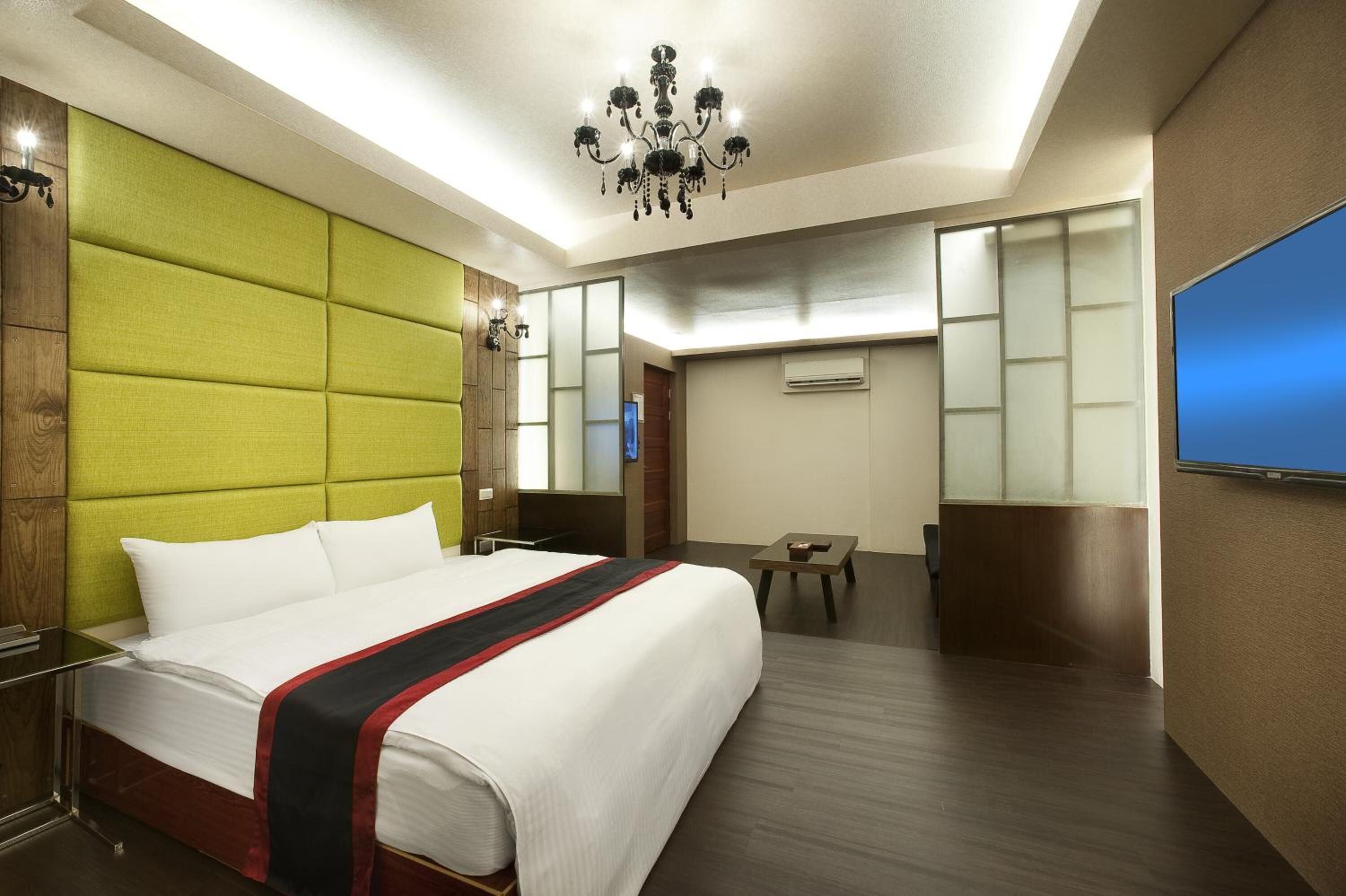 A hotel room with a large bed, a green headboard, a chandelier, a flat-screen TV, and a small table.