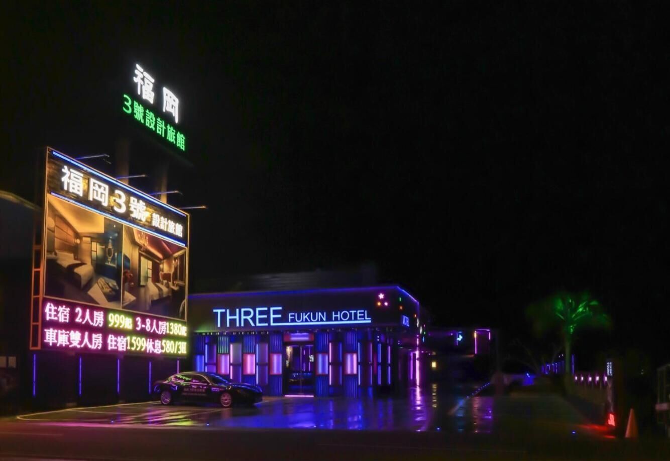 A hotel named 'THREE FUKUN HOTEL' with illuminated signage and a large billboard advertising rooms and prices.