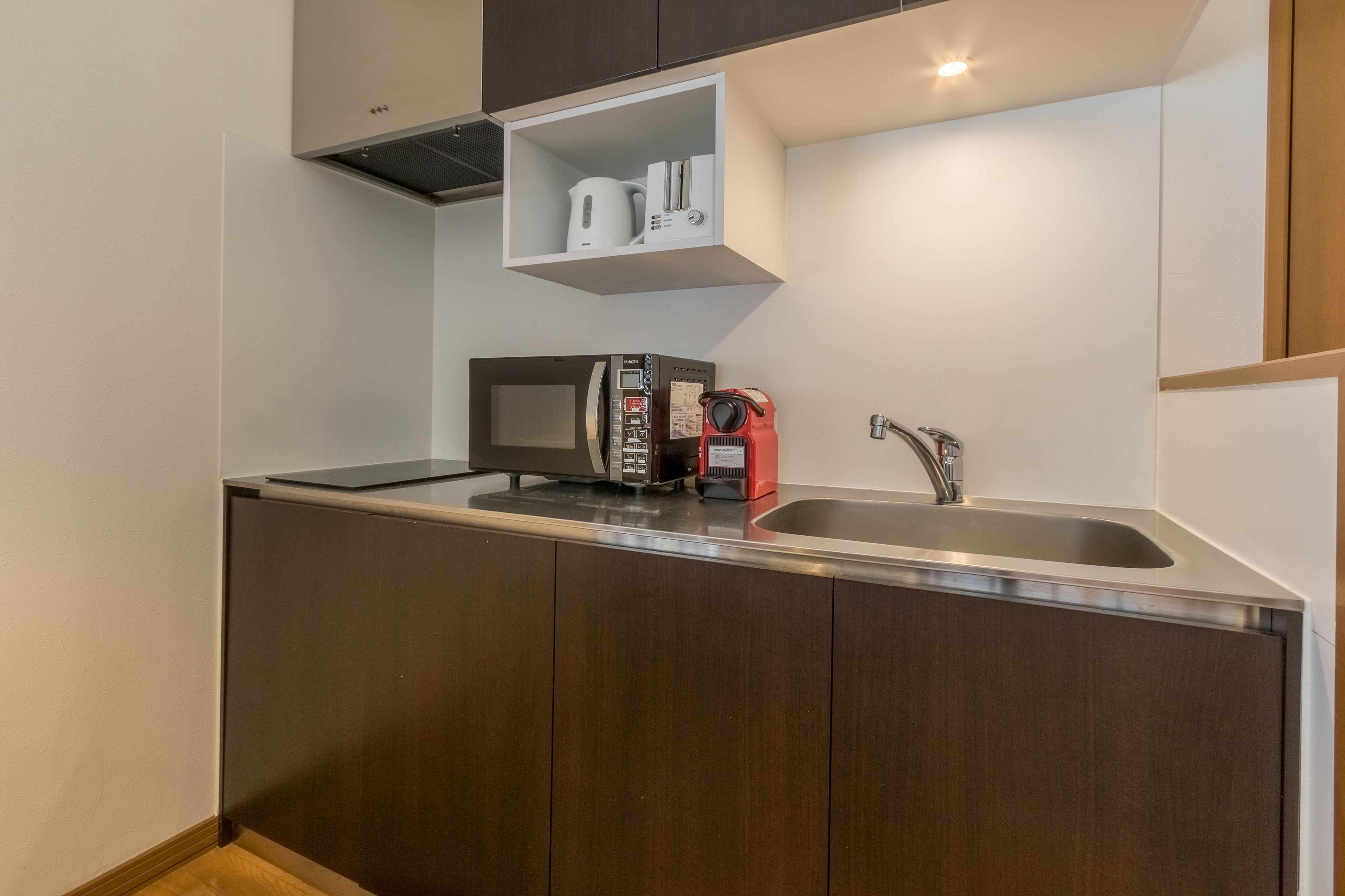 Una cocina compacta con armarios de madera oscura, un lavamanos de acero inoxidable, un microondas y una tetera roja.