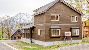 Un bâtiment en bois à deux étages, doté d’un toit en pente, avec une petite construction adjacente surmontée d’un toit rouge, et une montagne en arrière-plan.