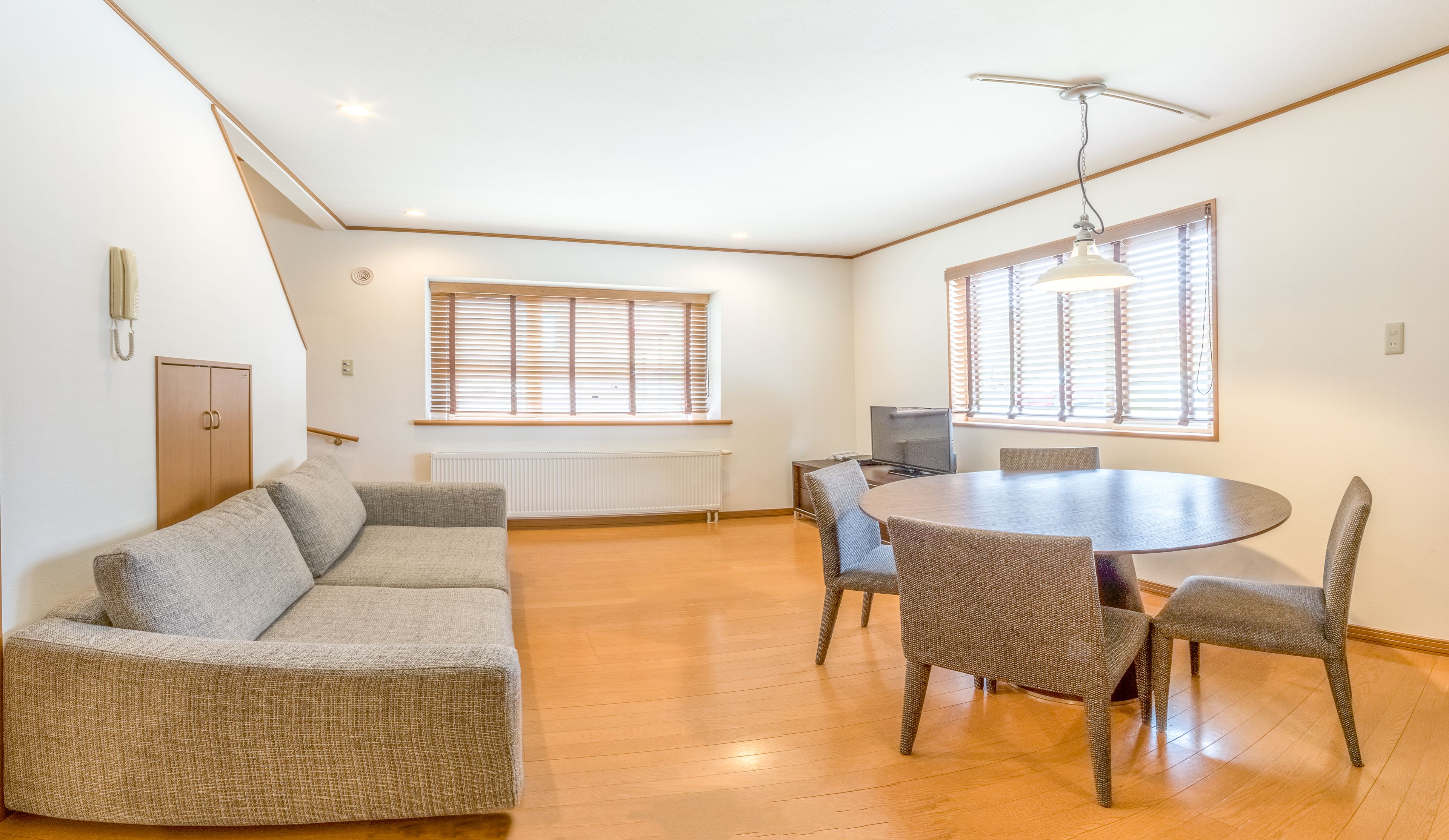 A spacious living room with a sofa, a dining table with chairs, and large windows with blinds.