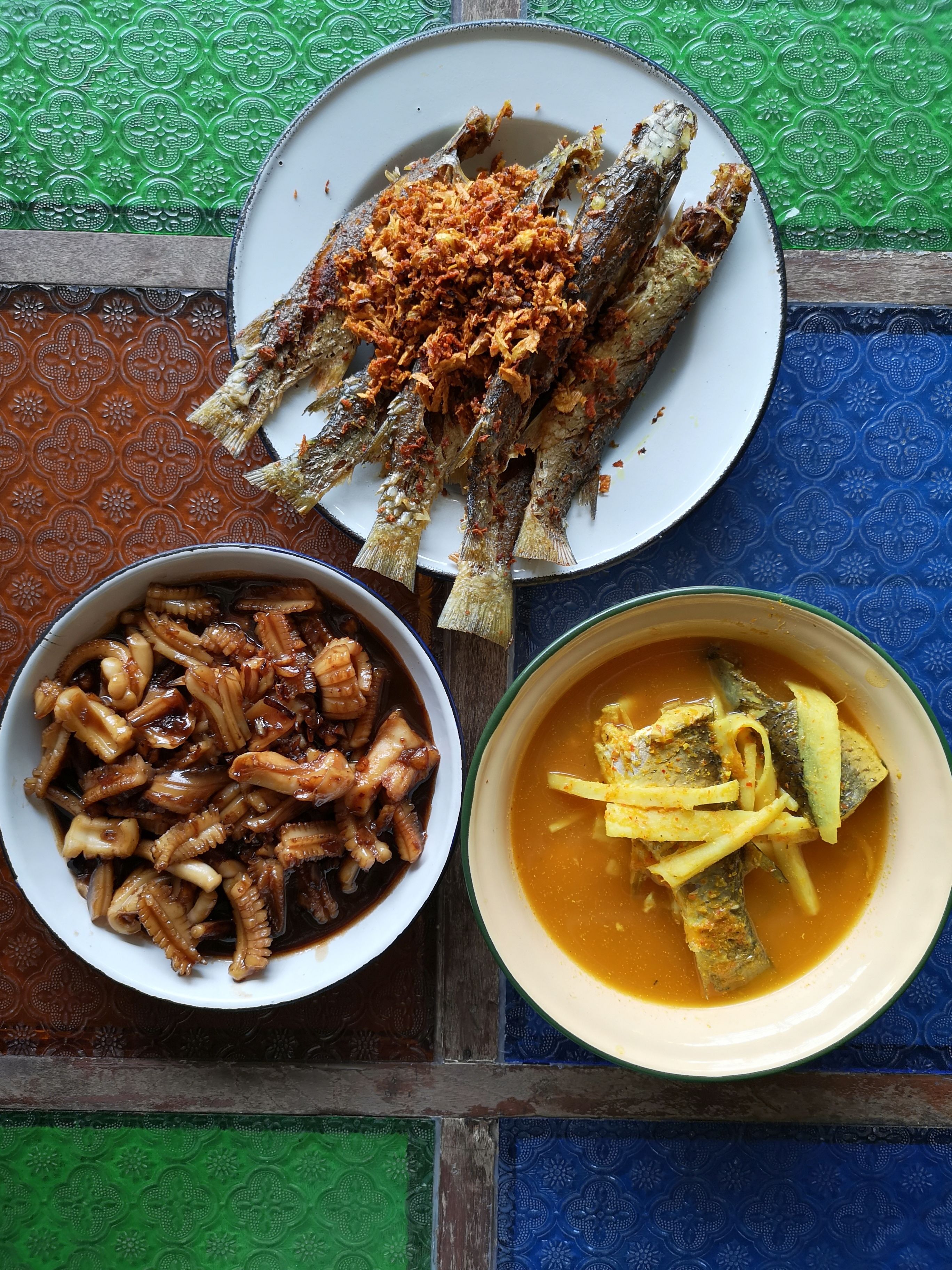 Ein Tisch mit einem Teller frittiertem Fisch, belegt mit knuspriger Gewürzmischung, einer Schale mit Oktopus und einer Schüssel Gemüsesuppe.
