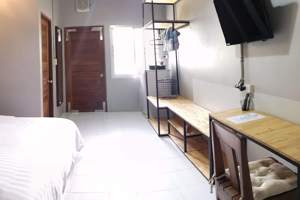 Un dormitorio con cama, escritorio de madera, silla, televisor y ventana.