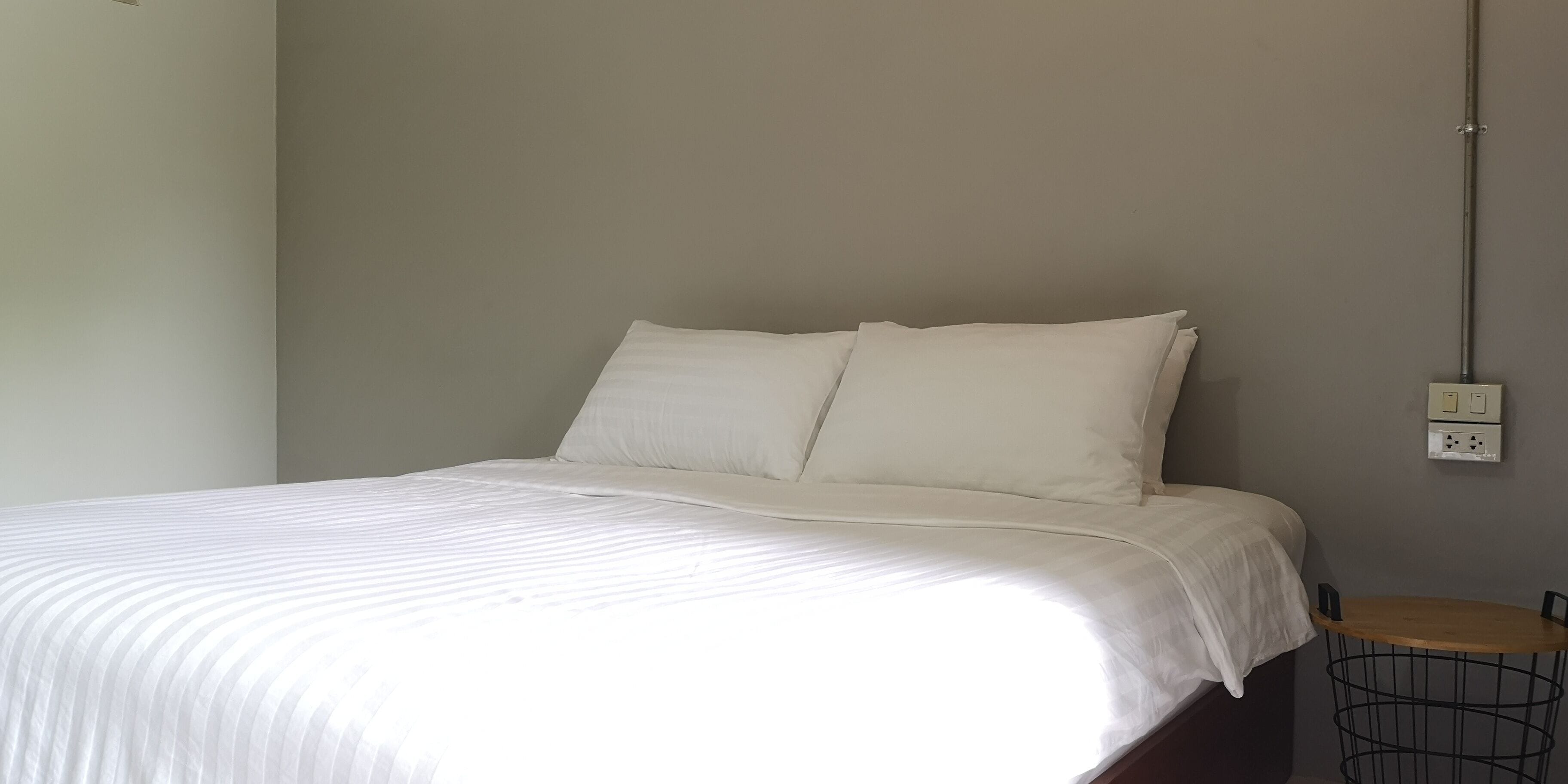 A neatly made bed with white linens in a room with a grey wall and a small wooden table with a metal basket beside it.