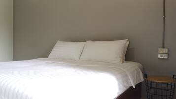 A neatly made bed with white linens in a room with a grey wall and a small wooden table with a metal basket beside it.