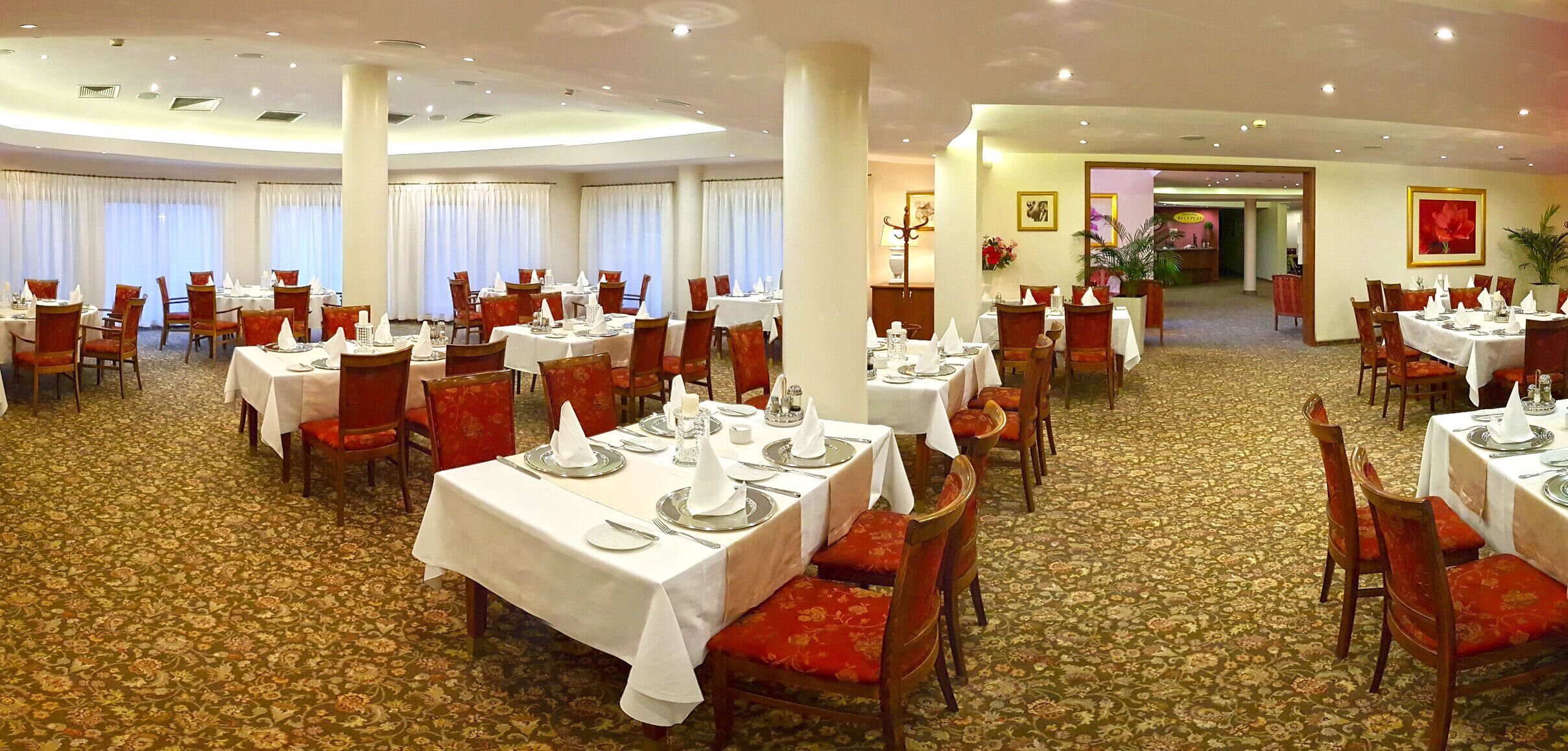 A hotel dining area with multiple tables set for dining, red upholstered chairs, and a carpeted floor.