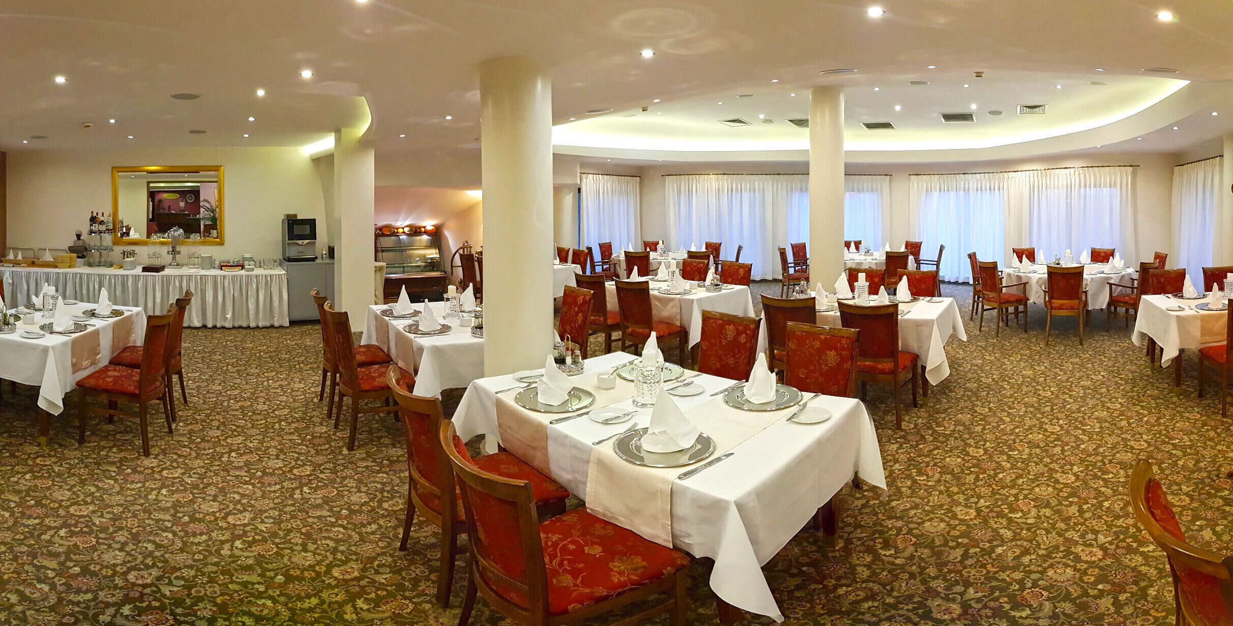 A hotel dining area with multiple tables set for service, carpeted flooring, and large windows with curtains.