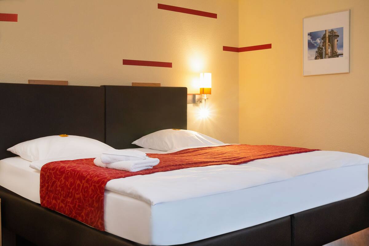 A neatly made bed with white linens and a red blanket, a wall-mounted light, and a framed picture on the wall.