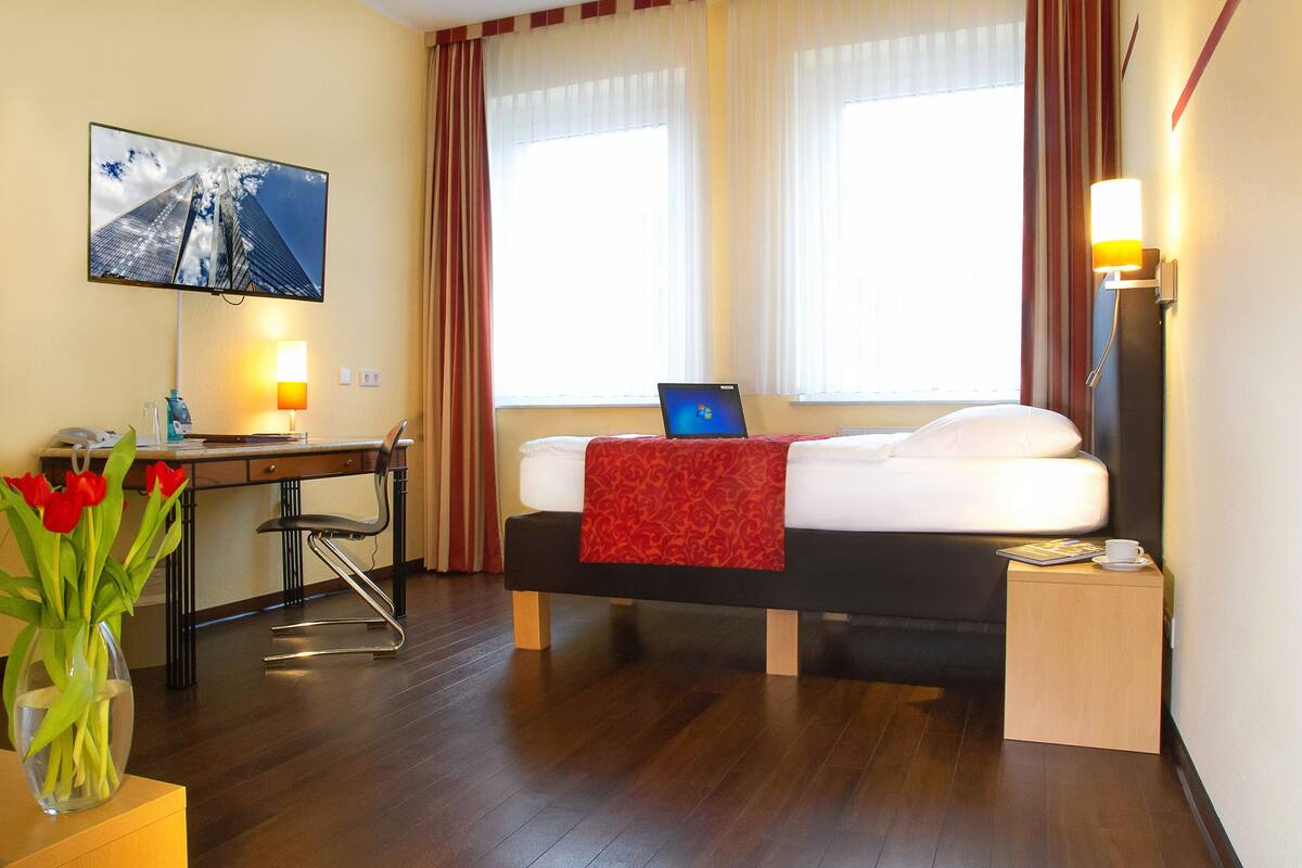 A hotel room with a bed, a desk with a laptop, a TV on the wall, and a vase with flowers.