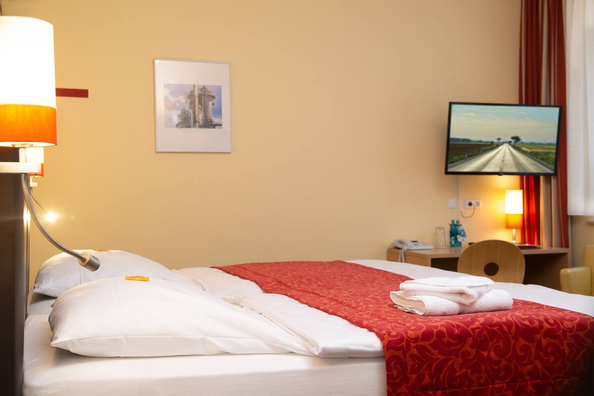A hotel room with a neatly made bed, a television on the wall, and a desk with a chair.