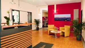 A reception area with a modern design, featuring a reception desk, two yellow armchairs, a small table with a vase of flowers, and a large cityscape painting on the wall. - Hotel NeuHaus (Dortmund)