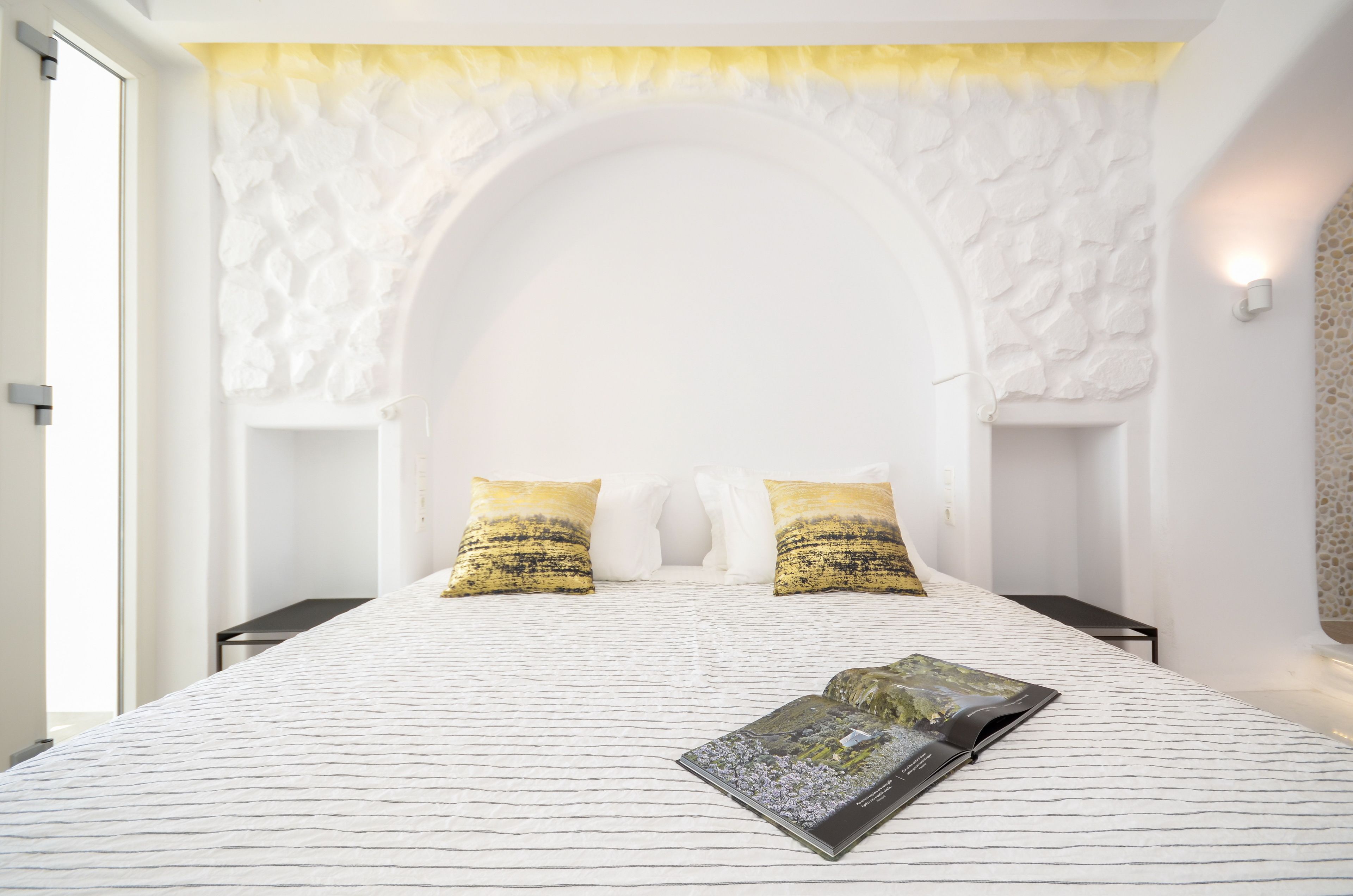Een slaapkamer met een witte, gestructureerde wand, een bed met gestreept beddengoed, twee kussens en een tijdschrift op het bed.