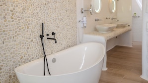 A modern bathroom with a freestanding bathtub, a large mirror, and a stone-textured wall. - Cyano Suites (Naxos)