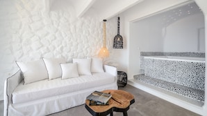 A modern living room with a white textured wall, a white sofa with multiple cushions, a wooden coffee table with books, a hanging light fixture, and a patterned bench against the wall. - Cyano Suites (Naxos)