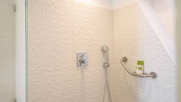 Zona de ducha con pared texturizada, un cabezal de ducha de mano, grifo empotrado en la pared y estante con artículos de aseo.