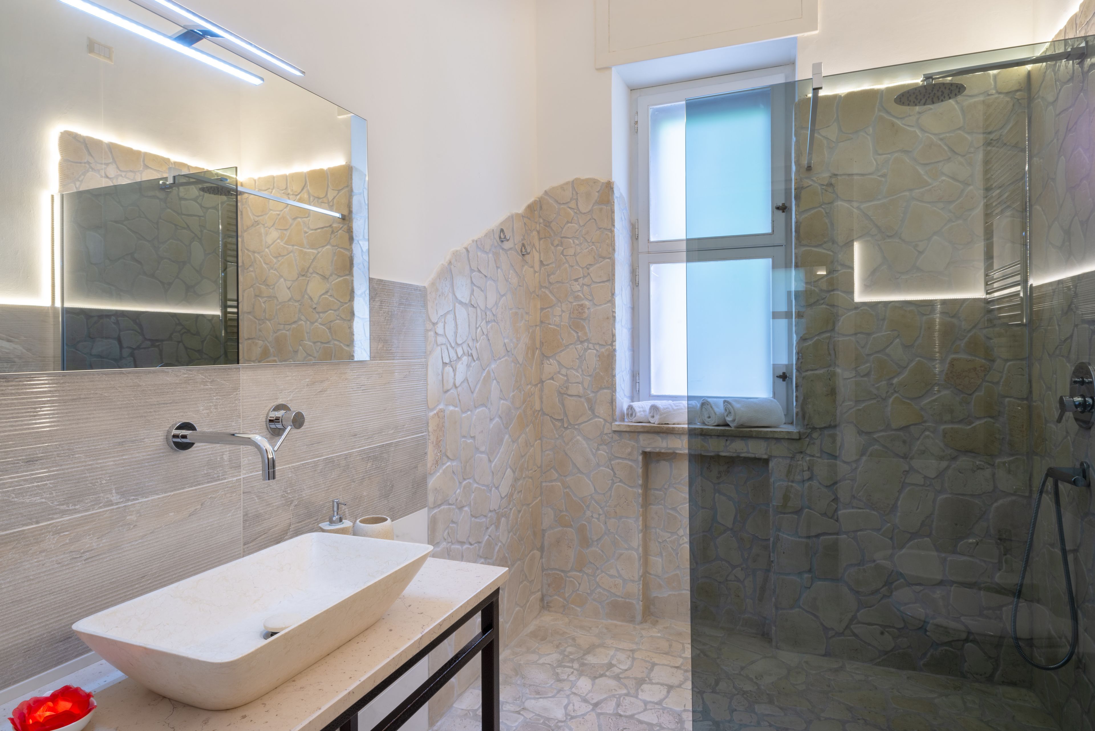 Ein Badezimmer mit Steinwand, einer gläsernen Dusche, einem Waschbecken und einem Fenster.