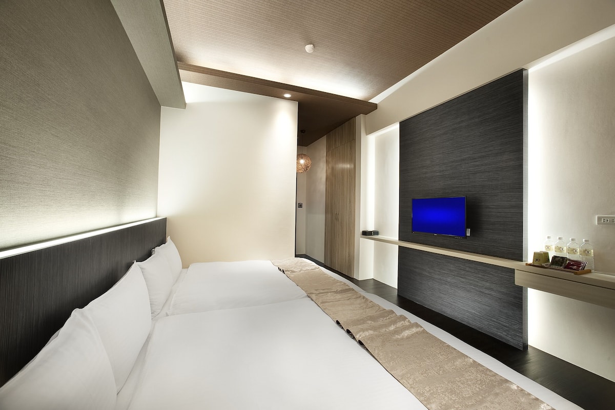 A modern hotel room with a large bed, a mounted TV, and a built-in shelf with toiletries.